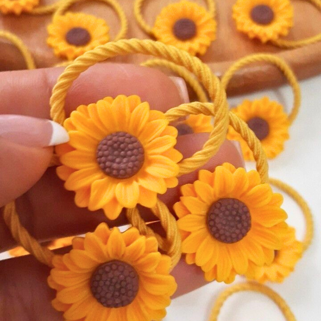 Sunflower-shaped hair ties with brown centers held by a hand against a blurred background of more hair ties.