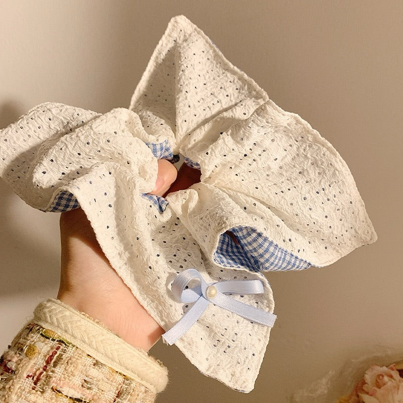 Hand holding a textured white fabric square shaped scrunchie with blue checkered interior and ribbon, against a neutral background.