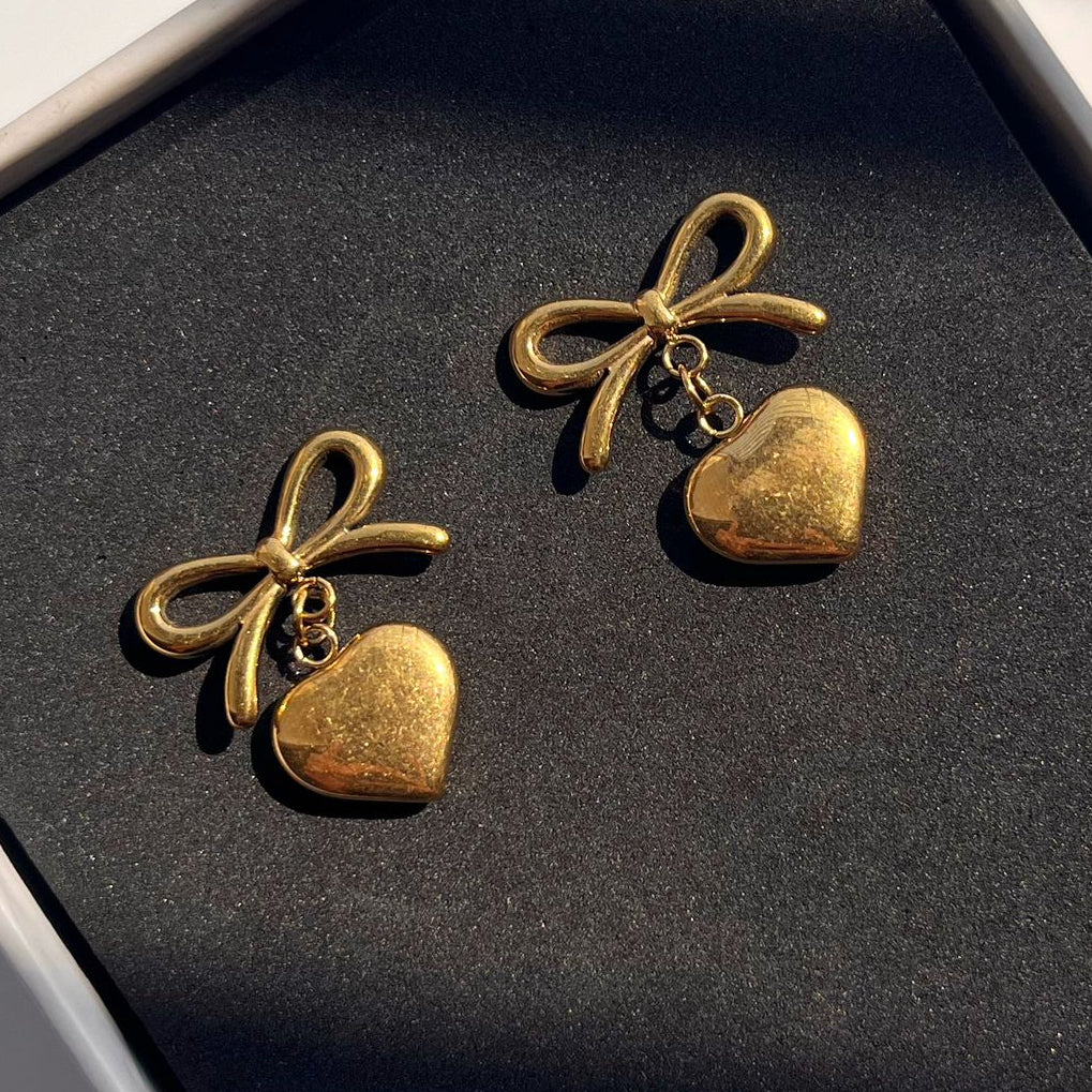 Pair of gold bow and heart-shaped earrings on a black box.