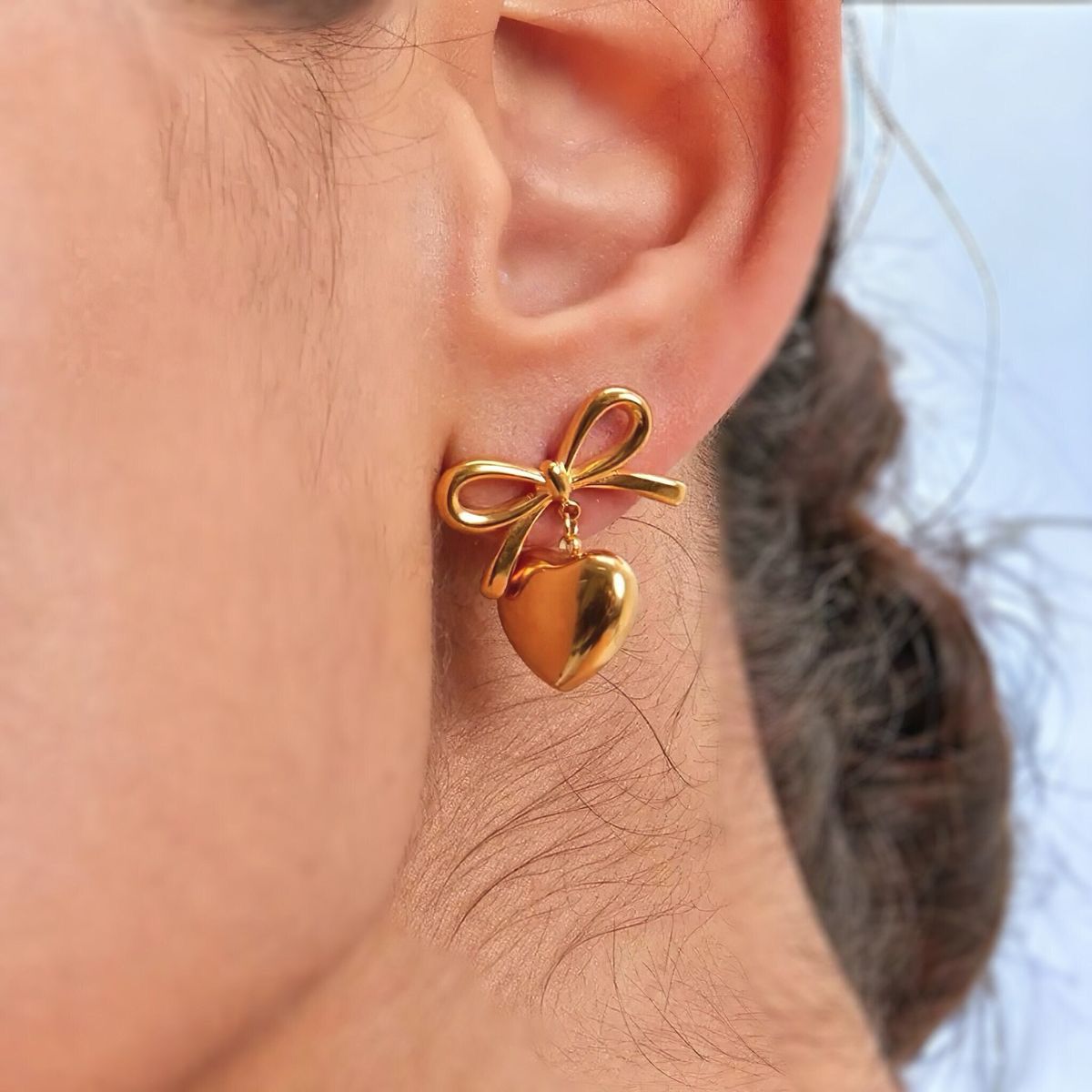 Gold bow and heart earring worn on an ear with a blurred background