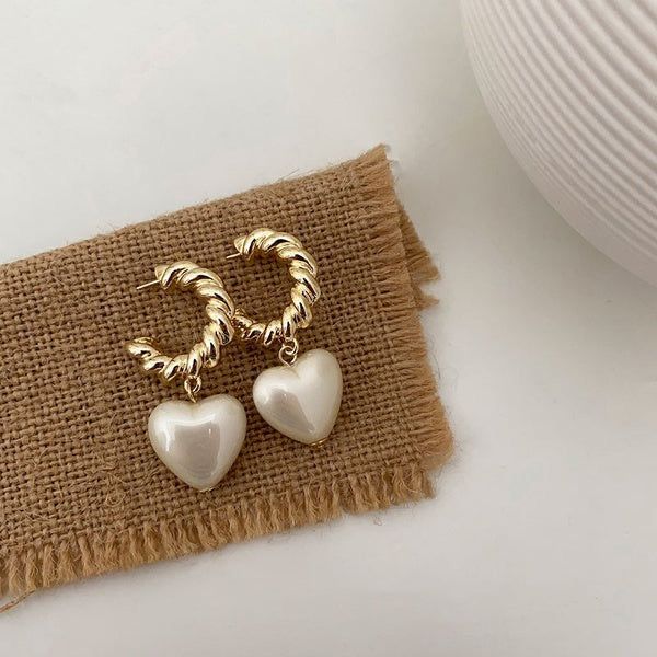 Gold hoop earrings with heart-shaped pearl charms on a textured fabric surface.