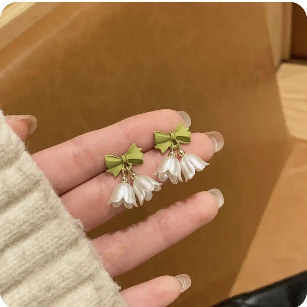 Hand holding a pair of earrings with green bows and white flowers against a brown background