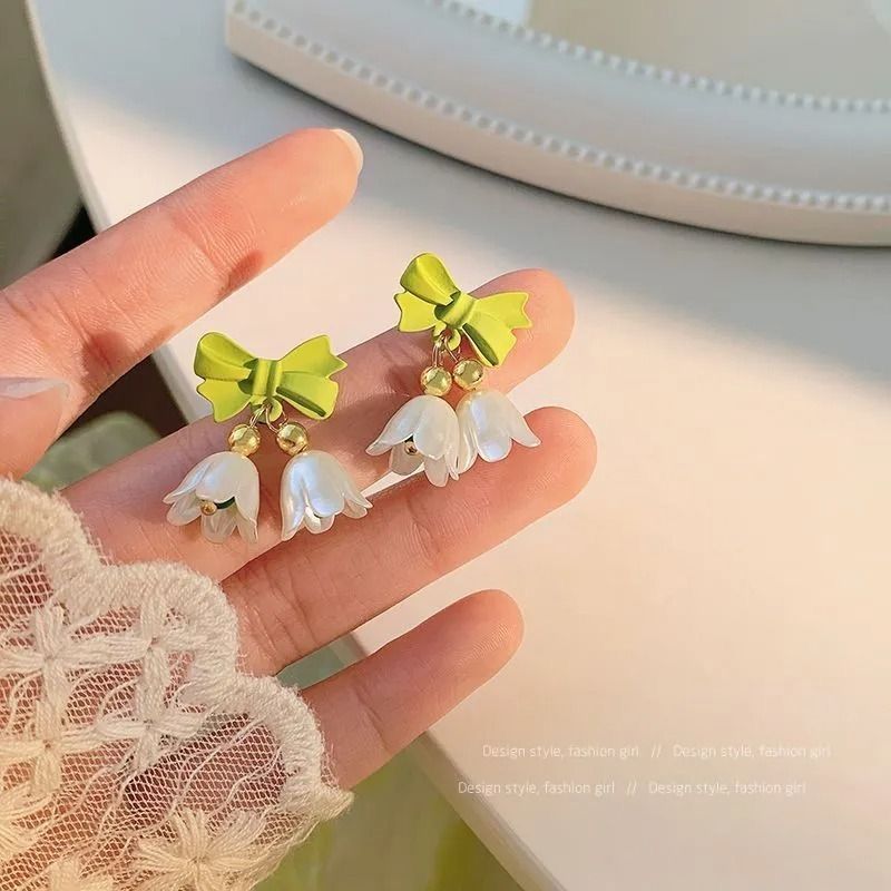 Green bow and white flower earrings held in a hand with a lace sleeve.