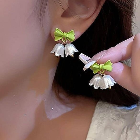 Earrings with white floral designs and green bows being held near an ear.