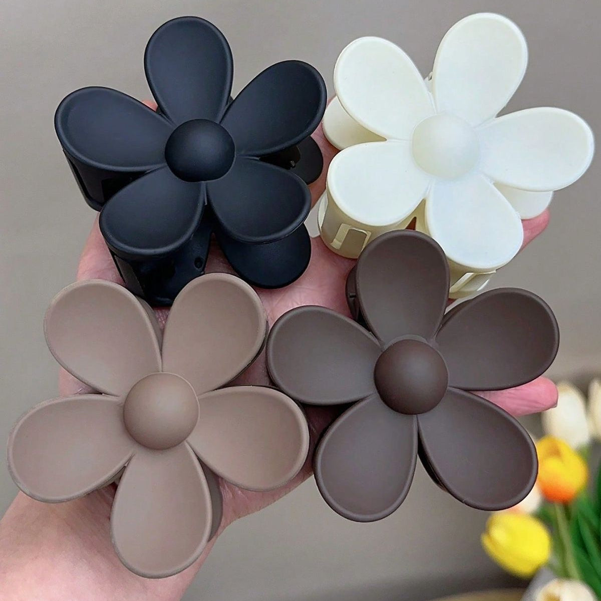 Four flower-shaped hair clips in black, white, beige, and brown held by a hand against a neutral background.