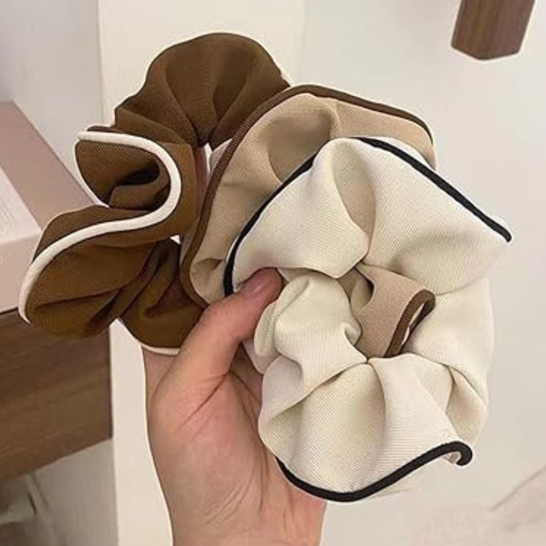 Three scrunchies in brown, beige, and white held by a hand against a neutral background.