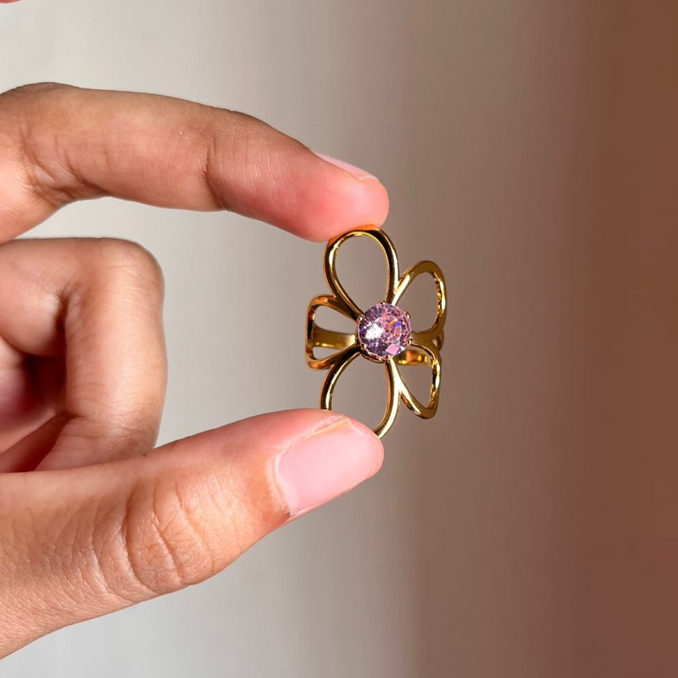 Gold flower-shaped ring with a pink gemstone held between fingers against a neutral background