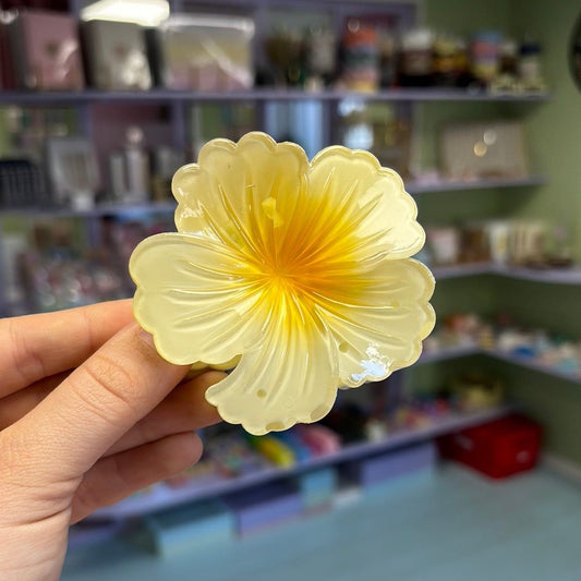 Hand holding a yellow flower-shaped hair clip with a blurred background of shelves.