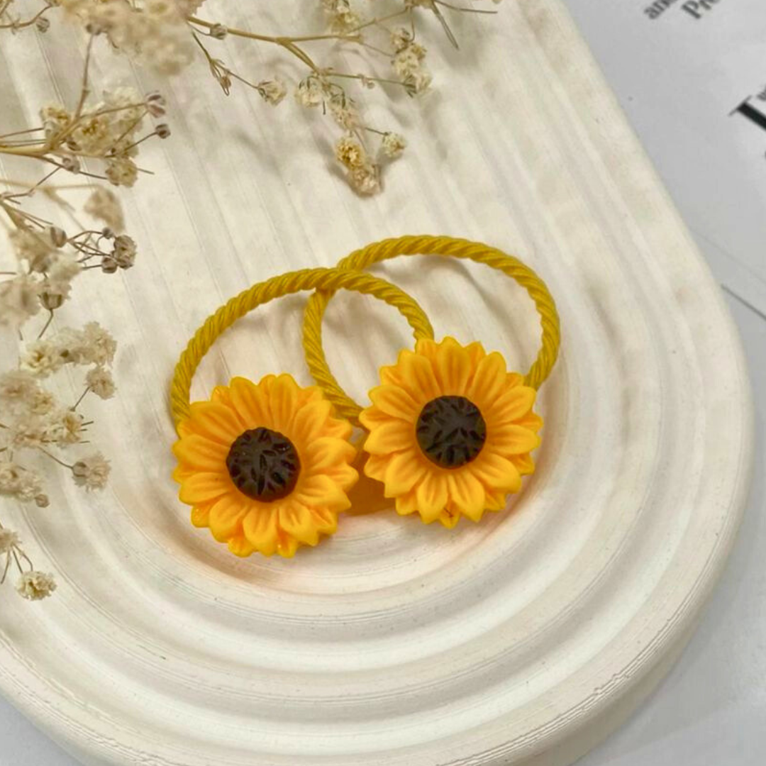 Two yellow hair ties with sunflower designs on a white surface with dried flowers.