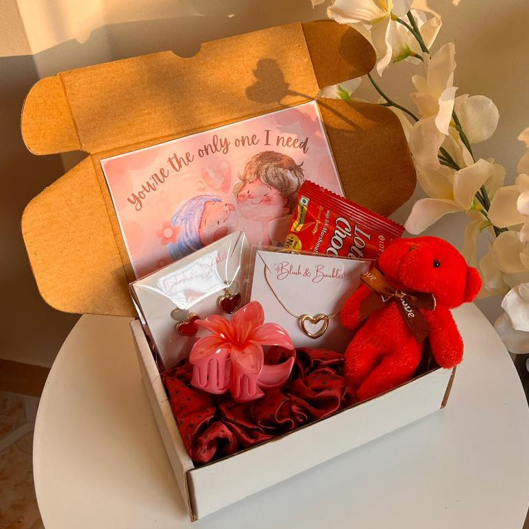 Gift box with a teddy bear, card, and other items on a table with flowers in the background