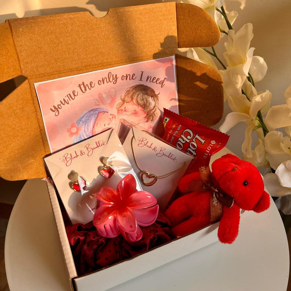 Gift box with a teddy bear, card, and other items on a white surface with flowers in the background.