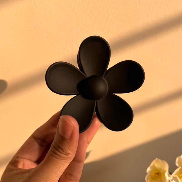 Black flower-shaped clip held by a hand on a beige background with a plant in the corner.