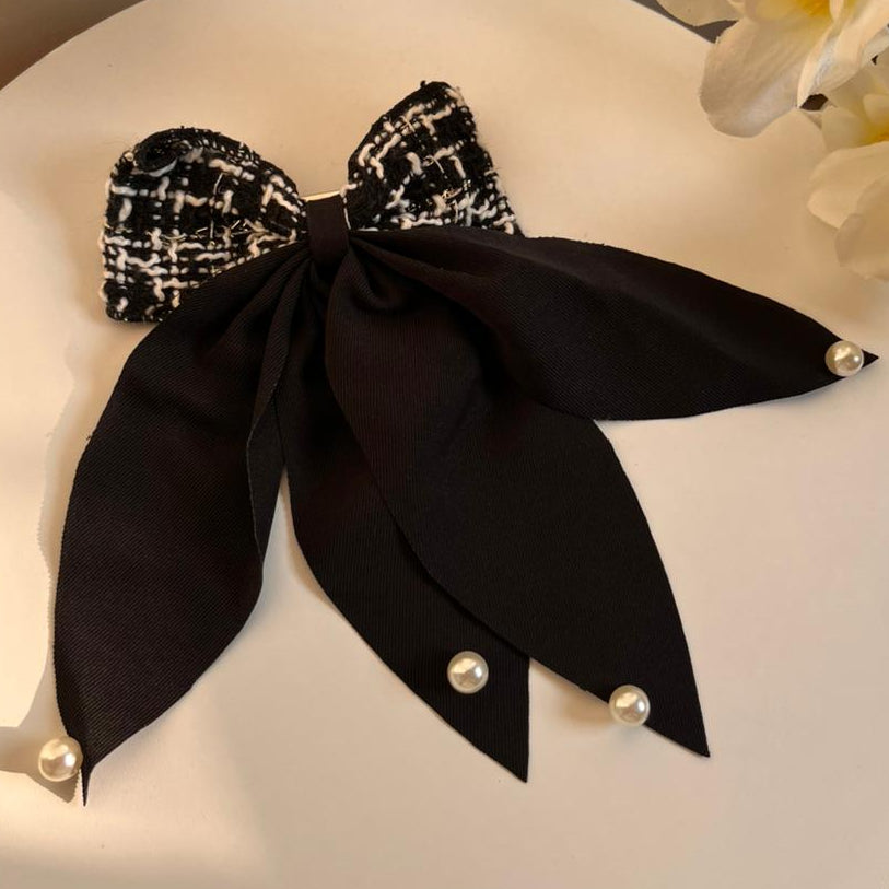 Black bow-shaped hair clip with white floral details on a white surface with flowers in the background