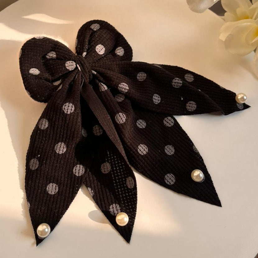 Black polka dot bow with pearl accents on a white surface with flowers in the background