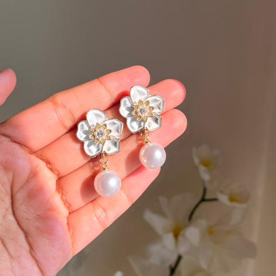 Hand holding a pair of floral earrings with pearl accents against a neutral background.