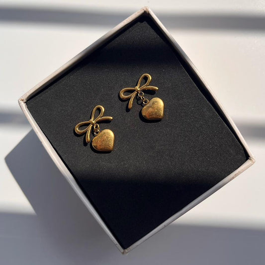 Gold bow and heart-shaped earrings in a black box on a white background