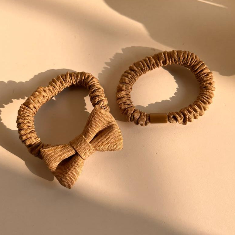 Two brown hair scrunchies with a bow on a beige surface