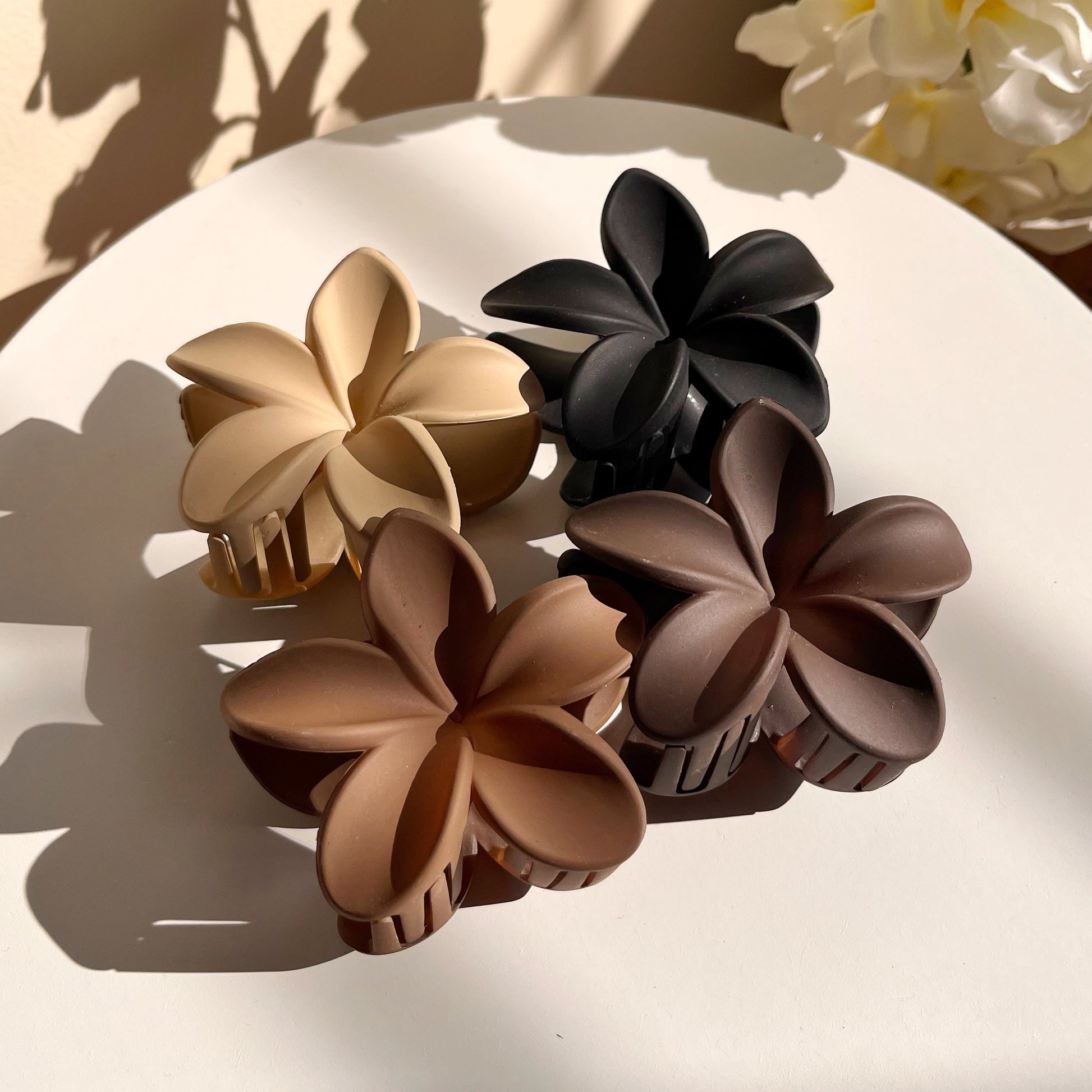 Set of four floral hair clips in beige, black, and brown on a white surface with a blurred background.