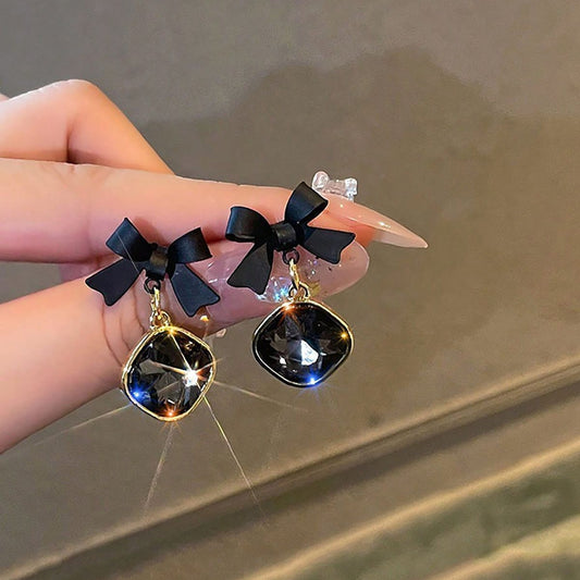 Black bow and crystal earrings held by a hand against a neutral background