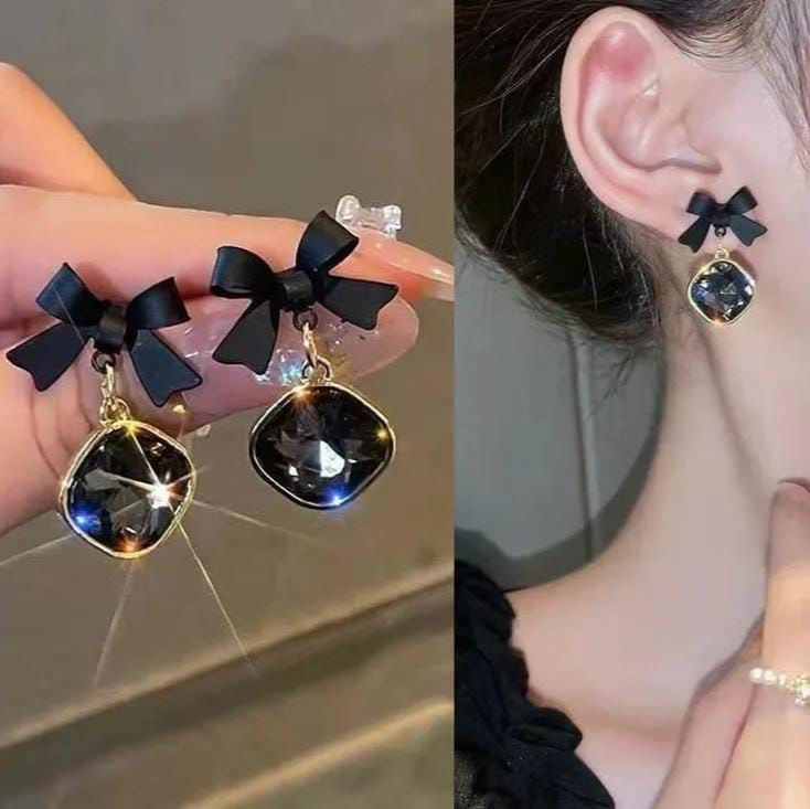 Black bow earrings with gold accents held by a hand and worn on an ear.