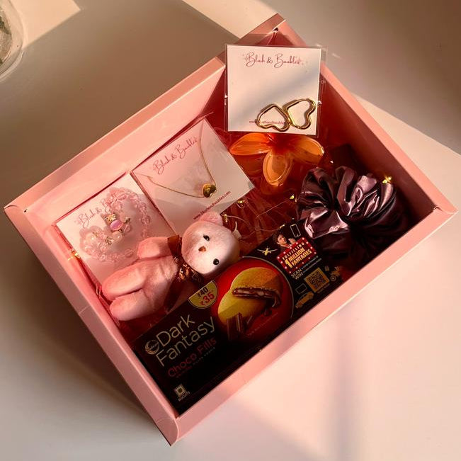 Gift box with various items including a jewellery, keychain, hair accessories and Chocolate on a light surface.