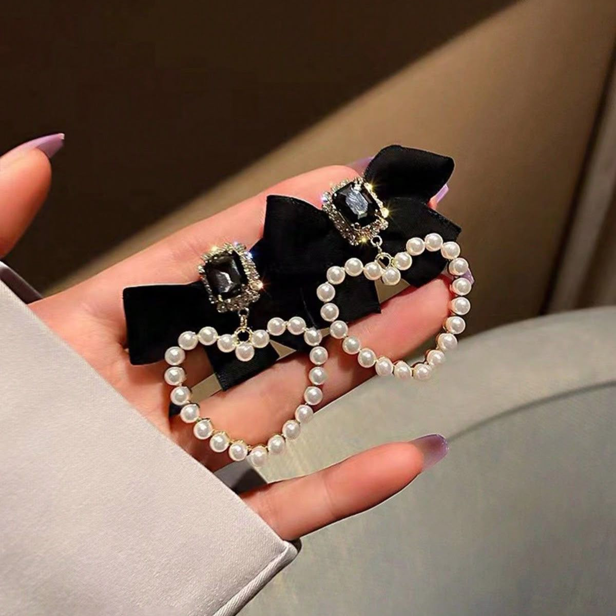 Hand holding two decorative heart shaped earrings with a satin bow and pearl detailing.