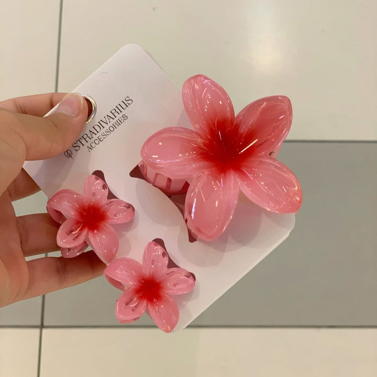 Pink flower-shaped hair clips on a white card held by a hand against a tiled floor background.