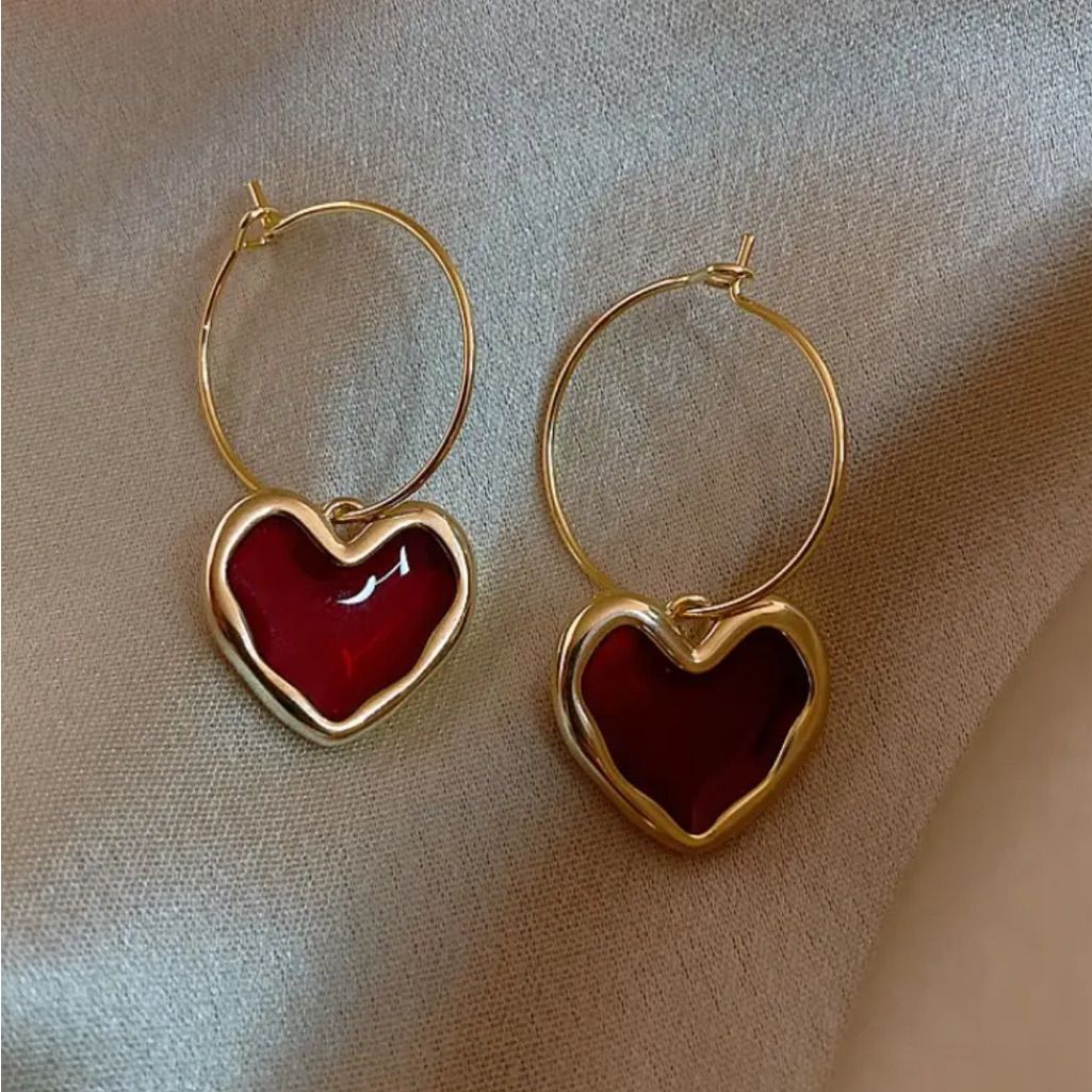 Gold hoop earrings with red heart charms on a beige fabric background