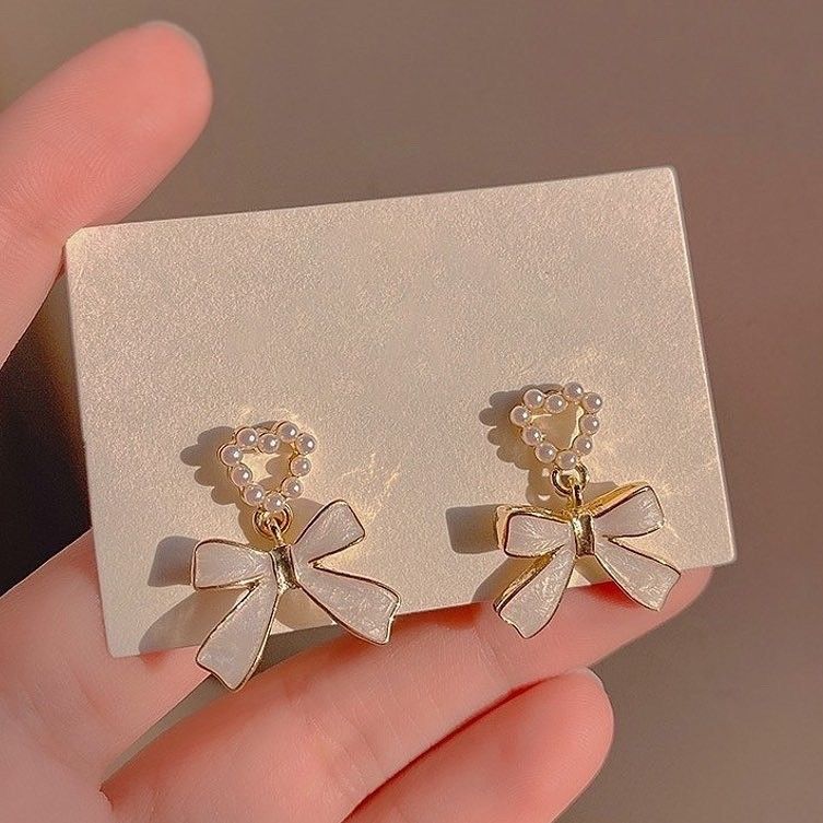 Gold bow-shaped earrings with pearl details on a beige card held by a hand.