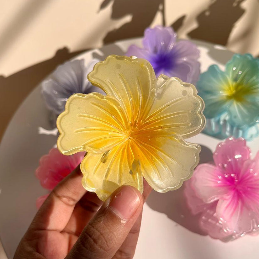 Hand holding a yellow flower-shaped Hair Claw Clip with colorful flowers in the background.