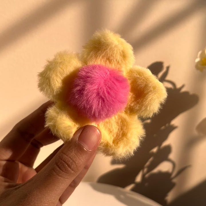 Hand holding a yellow flower-shaped hair clip with a pink centre against a beige background